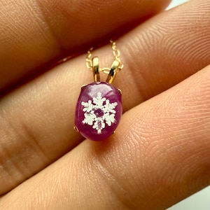 Könnte beinhalten: Eine goldene Halskette mit einem Anhänger. Der Anhänger hat einen tiefroten, oval geformten Stein mit einem weißen Schneeflocken-Design. Der Anhänger hängt an einer feinen Goldkette und ist ein elegantes und festliches Accessoire.