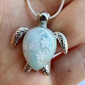 Può includere: Ciondolo a forma di tartaruga argentata con guscio azzurro chiaro e bianco, decorato con motivi a fiocco di neve. Il ciondolo è attaccato a una delicata catena d'argento. Le pinne e la testa della tartaruga hanno un aspetto strutturato.