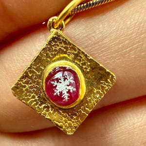 Könnte beinhalten: Goldene Halskette mit Anhänger. Der rautenförmige Anhänger hat eine strukturierte Oberfläche und einen roten, runden Edelstein mit weißem Schneeflockenmuster. Die Kette ist ebenfalls aus Gold.