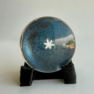 Peut inclure: Un presse-papier sphérique avec un intérieur bleu profond et scintillant, orné d'un flocon de neige blanc en son centre. La sphère en verre transparent repose sur un support noir texturé à trois branches qui maintient l'objet en place.