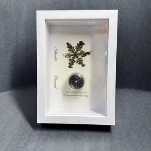 Peut inclure: Présentation encadrée blanche avec un flocon de neige préservé et un petit objet rond. Le flocon de neige est une silhouette marron foncé détaillée. Le texte "Silhouette Preserved" est à gauche, et "Every snowflake holds a story" est en dessous.