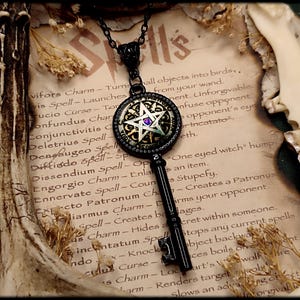 May include: Black key pendant necklace with a circular pendant featuring a pentagram and a purple gem. The key rests on a parchment with the word "Spells" and spell names. Dried flowers and a bone are also present.