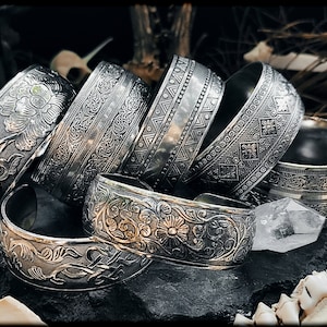 Pode incluir: Uma coleção de pulseiras de metal prateado com intrincados desenhos florais e geométricos gravados. As pulseiras estão empilhadas umas sobre as outras, criando uma exibição visualmente atraente.