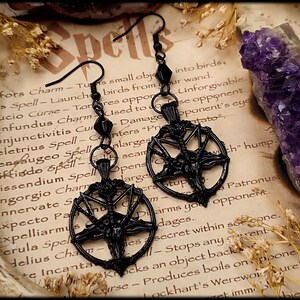 Può includere: Orecchini a pentagramma neri dal design gotico. Gli orecchini presentano un ciondolo a pentagramma dettagliato con una figura centrale, sospeso a una catena di perline. Sono esposti su uno sfondo di carta vintage con testo.