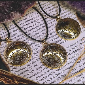Pode incluir: Três pingentes de metal dourado com cordões pretos. Cada pingente apresenta um símbolo e uma inscrição diferentes, incluindo "DONAY", "AGIOS" e "OTHEO".
