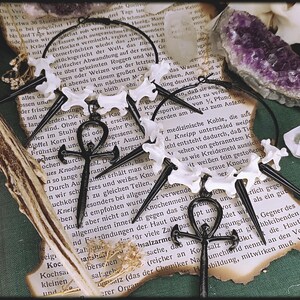 Könnte beinhalten: Ein Paar schwarze und weiße Ohrringe mit einem Ankh-Symbol-Anhänger. Die Ohrringe sind aus Knochen und Metall gefertigt und haben einen gotischen Stil.