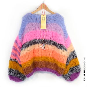 May include: A colorful, oversized sweater with a striped design in shades of purple, pink, orange, yellow, and black. The sweater has long sleeves and a round neckline. A tag with the letters "MYPZ" hangs from the sweater.