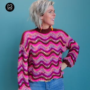 Op de afbeelding: Een roze, paarse en bruine gebreide trui met een golvend patroon. De trui heeft een hoge hals en lange mouwen.