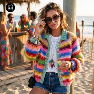 Op de afbeelding: Een kleurrijk, gestreept vest met een groene kraag en roze knopen. De trui heeft een regenboog aan kleuren en wordt gecombineerd met een spijkerbroek. De foto is buiten genomen, met een strand en een bar op de achtergrond.