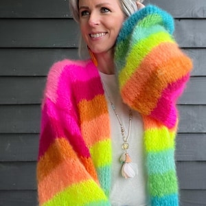 Op de afbeelding: Een regenboog gestreepte, pluche cardigan met lange mouwen. De cardigan is open en de persoon die hem draagt ​​lacht.