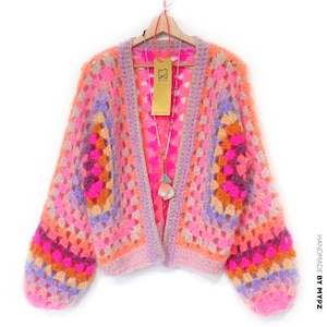 Könnte beinhalten: Ein farbenfroher, offener Häkelcardigan in Rosa-, Orange- und Lilatönen. Das handgefertigte Kleidungsstück hat ein Granny-Square-Design mit langen Ärmeln und einem weichen, strukturierten Aussehen. Ein Etikett mit dem Markennamen hängt am Ausschnitt.