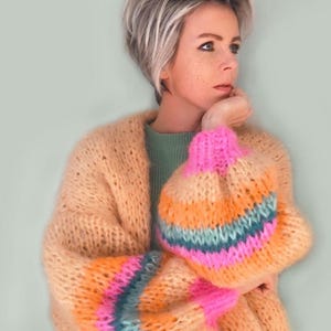 Op de afbeelding: Een licht perzikkleurige gebreide cardigan met lange mouwen en een gestreept patroon in roze, oranje, teal en blauw. De cardigan is gemaakt van een zacht, pluche garen.
