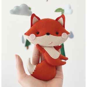 May include: A red felt fox with a white belly and a fluffy tail. The fox has black eyes and pink cheeks. The fox is a handmade toy and is part of a mobile.