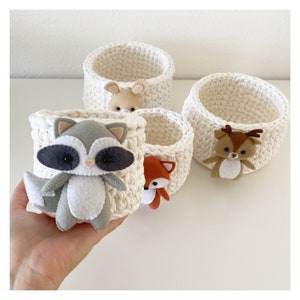 May include: Four white crocheted baskets with animal-shaped felt embellishments. The baskets are filled with felt animals, including a raccoon, a fox, a bunny, and a deer.