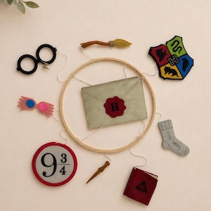 May include: A circular mobile featuring felt cutouts of iconic items from a fantasy book series. Includes glasses, a broom, a crest, an envelope, a sock with text, a wand, a book, and a platform sign. The items are in various colors, including black, red, and gold.