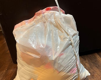 May include: A large, clear plastic bag filled with items, possibly clothing or textiles. A white measuring tape hangs down the front of the bag, showing measurements in inches. The bag is sitting on a dark wood floor.