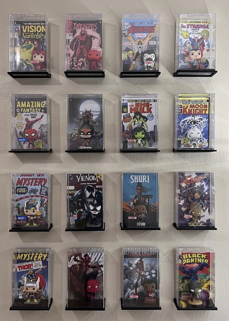 May include: A wall display of comic books in clear plastic cases with small figurines of characters from the comics. The comics include titles like "Vision", "Avengers", "Amazing Fantasy", "She-Hulk", "Moon Knight", "Journey into Mystery", "Venom", "Shuri", "Ultimate Fallout", and "Black Panther".