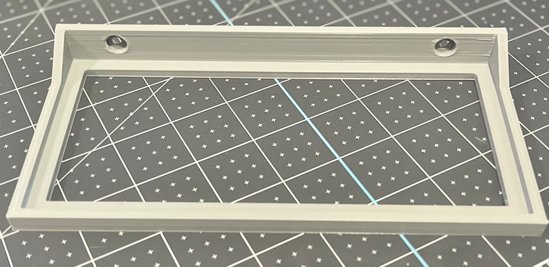 May include: A gray plastic frame with two holes on the top. The frame is designed to hold a small object, such as a picture or a piece of paper.