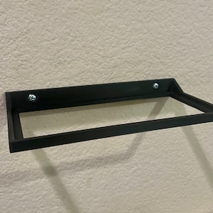 May include: A black metal shelf with a rectangular shape, mounted to a wall with two screws. The shelf has a simple design and is ideal for displaying small items.