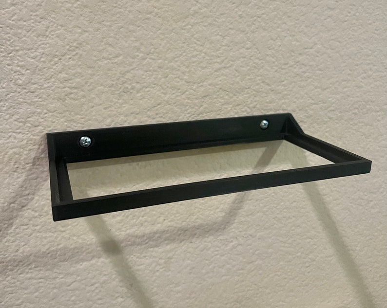 May include: A black metal shelf with a rectangular shape, mounted to a wall with two screws. The shelf has a simple design and is ideal for displaying small items.