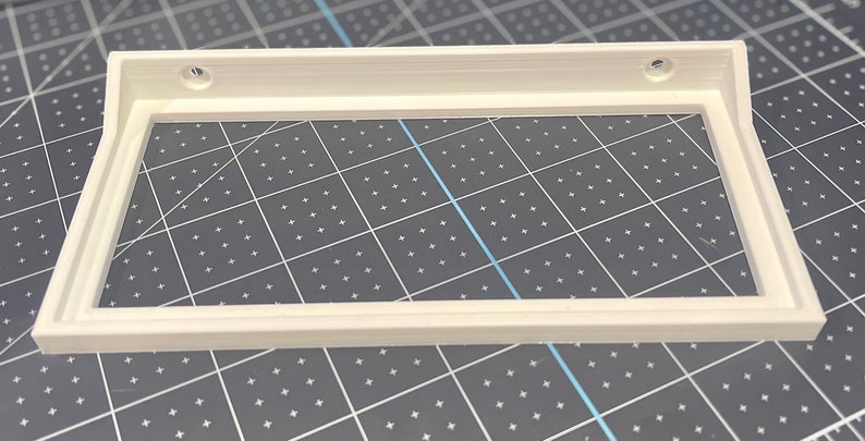 May include: White plastic frame with two screw holes on the top edge. The frame is designed to hold a picture or other artwork.