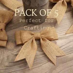 May include: A pack of five tan burlap bows with a woven texture and pointed ends. The bows are arranged around a gray metal container holding green foliage. The text PACK OF 5 Perfect for Crafting is printed in white over the center.