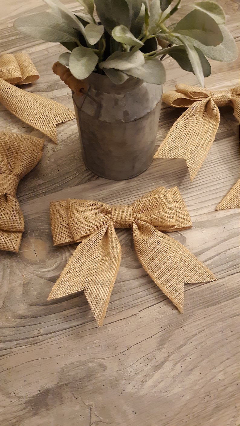 May include: Several tan burlap bows with a woven texture and pointed ends are arranged around a gray metal pitcher containing green foliage. The bows feature a central knot and two symmetrical loops with long, dangling tails.