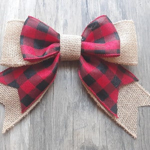 May include: A decorative bow featuring a combination of natural tan burlap and a red and black buffalo plaid fabric. The bow has a central burlap knot, two top loops, and two bottom tails with angled ends, showcasing a layered texture design.