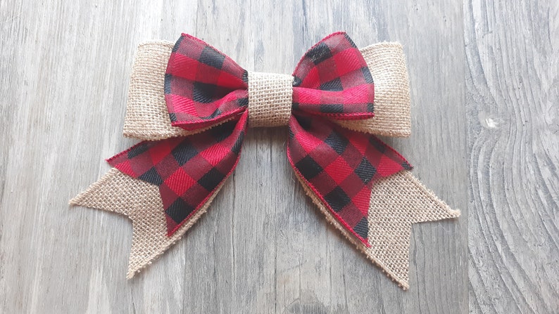 May include: A decorative bow featuring a combination of natural tan burlap and a red and black buffalo plaid fabric. The bow has a central burlap knot, two top loops, and two bottom tails with angled ends, showcasing a layered texture design.