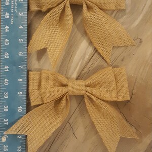 May include: Two tan burlap bows with a woven texture, each measuring approximately 5 inches in width, positioned next to a blue Empire Level T-square ruler that displays both imperial and metric measurement markings clearly.