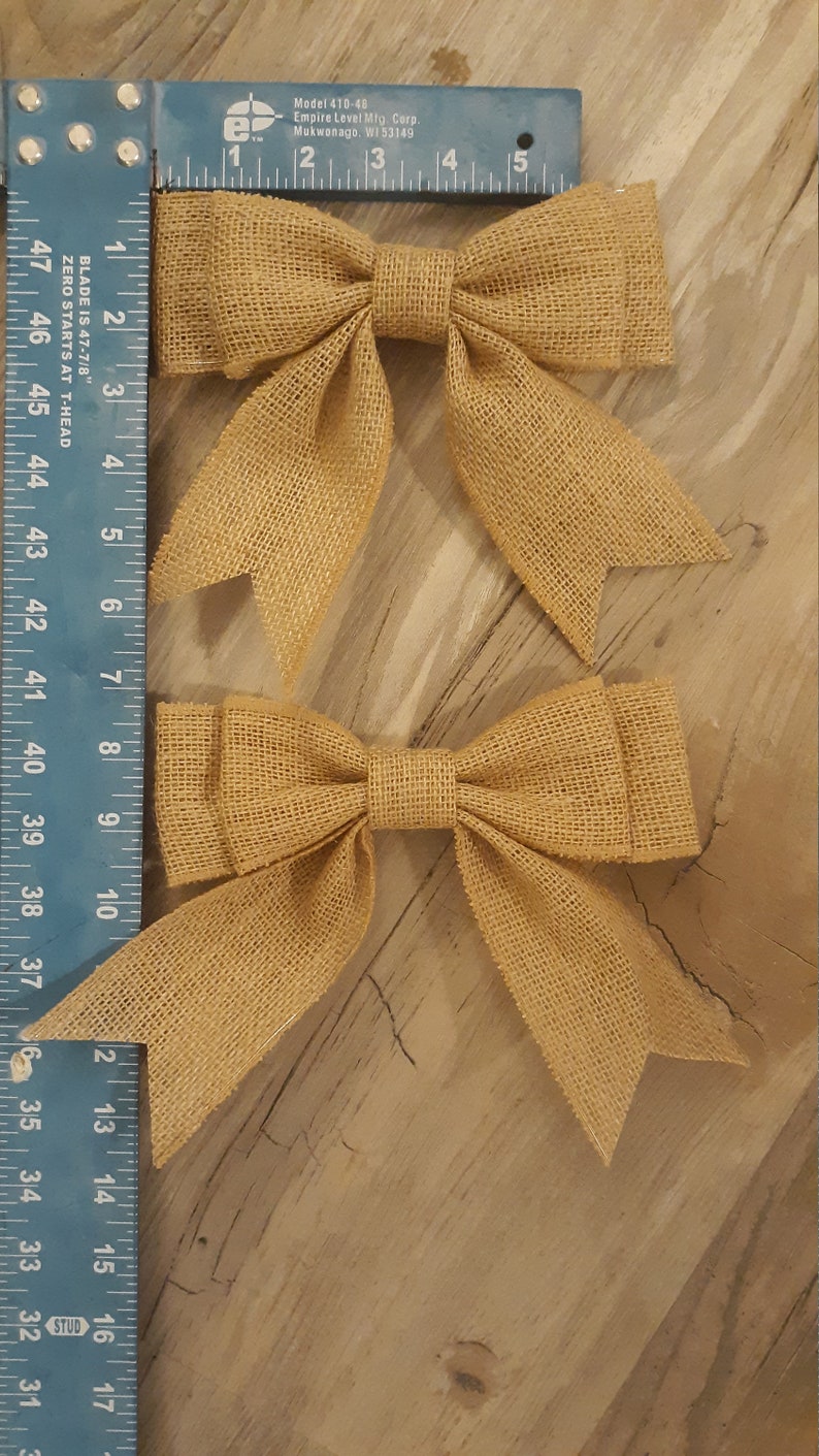 May include: Two tan burlap bows with a woven texture, each measuring approximately 5 inches in width, positioned next to a blue Empire Level T-square ruler that displays both imperial and metric measurement markings clearly.