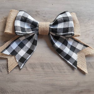 May include: A decorative bow featuring a black and white buffalo check pattern layered over tan burlap fabric. The center is cinched with a matching tan burlap band, and the ends of the bow are cut into pointed tails with frayed edges.