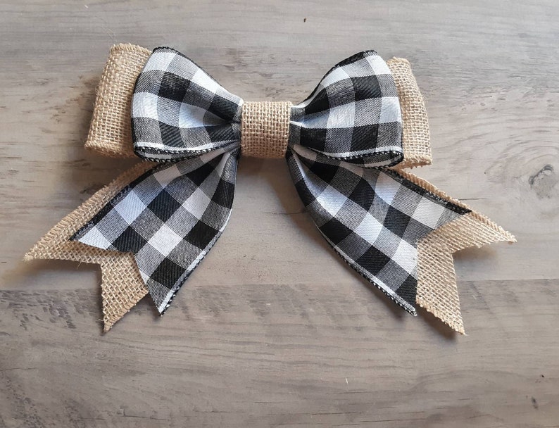 May include: A decorative bow featuring a black and white buffalo check pattern layered over tan burlap fabric. The center is cinched with a matching tan burlap band, and the ends of the bow are cut into pointed tails with frayed edges.