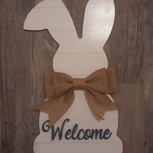 May include: A white wooden wall hanging shaped like a rabbit silhouette with horizontal panel lines. The rabbit features a tan burlap bow tied at the neck and the word Welcome in dark blue script lettering attached to the lower section.