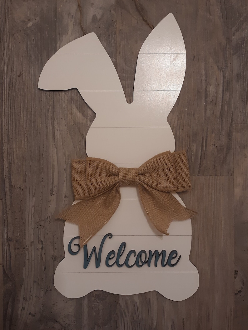 May include: A white wooden wall hanging shaped like a rabbit silhouette with horizontal panel lines. The rabbit features a tan burlap bow tied at the neck and the word Welcome in dark blue script lettering attached to the lower section.