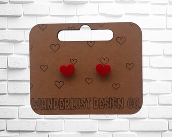 May include: A pair of small, solid red heart-shaped stud earrings attached to a brown rectangular display card. The card features a repeating pattern of small black heart outlines and the text WANDERLUST DESIGN CO. printed at the bottom.