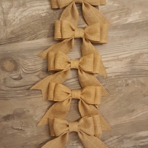 May include: A vertical arrangement of five tan burlap bows. Each bow features a central knot and two downward-pointing tails with V-shaped notched ends. The woven texture of the natural fiber material is visible across all five decorative bows.