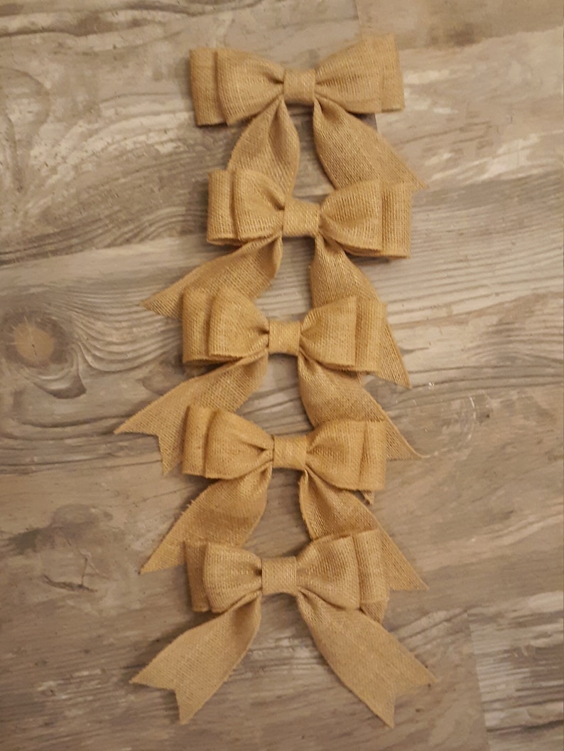 May include: A vertical arrangement of five tan burlap bows. Each bow features a central knot and two downward-pointing tails with V-shaped notched ends. The woven texture of the natural fiber material is visible across all five decorative bows.