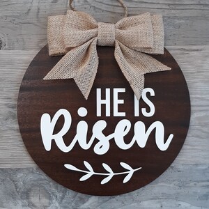 May include: A circular dark brown wooden sign featuring the white text HE IS Risen and a white leaf graphic. The sign is topped with a tan burlap bow and includes a jute twine loop for hanging, creating a decorative wall piece for display.