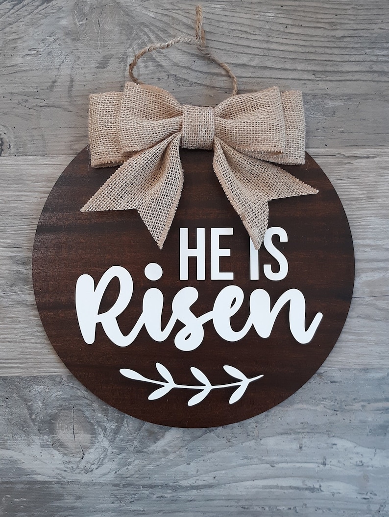 May include: A circular dark brown wooden sign featuring the white text HE IS Risen and a white leaf graphic. The sign is topped with a tan burlap bow and includes a jute twine loop for hanging, creating a decorative wall piece for display.