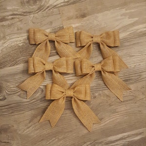May include: A set of five tan burlap bows, each featuring a central knot and two downward-pointing tails with V-shaped notched ends. The bows are constructed from a coarse, woven fabric with a visible texture and a consistent light brown color.