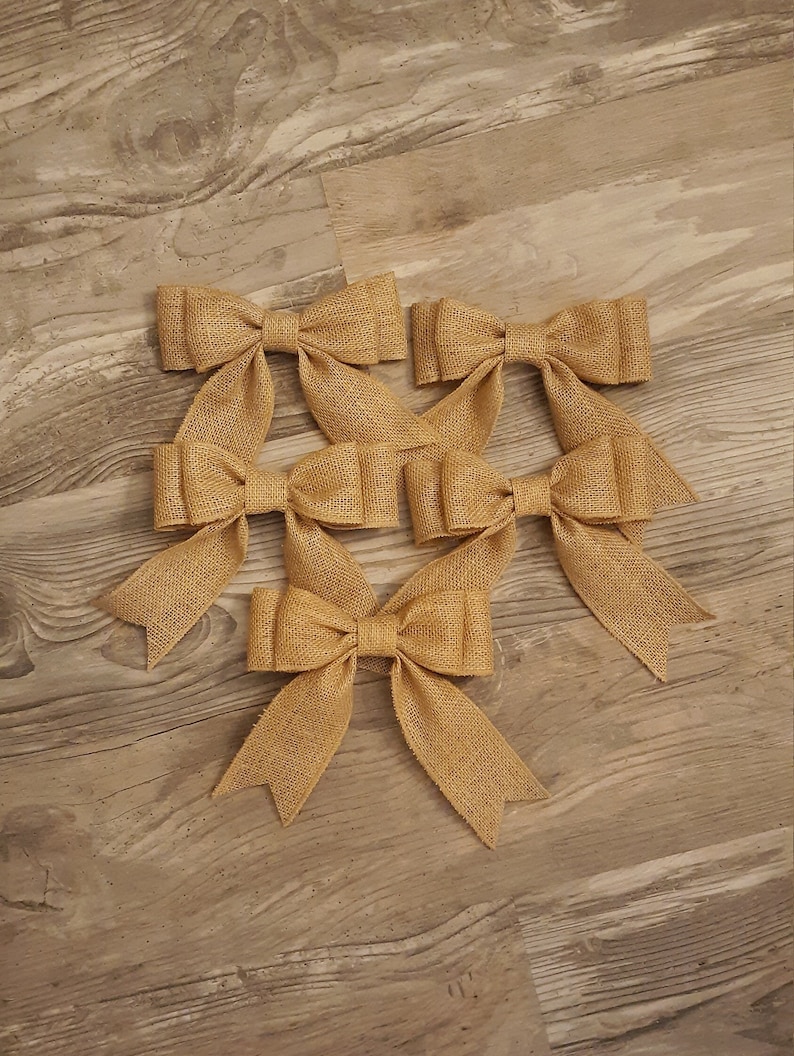 May include: A set of five tan burlap bows, each featuring a central knot and two downward-pointing tails with V-shaped notched ends. The bows are constructed from a coarse, woven fabric with a visible texture and a consistent light brown color.