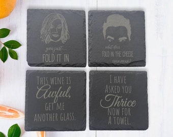 May include: A set of four square dark gray slate coasters with engraved text and illustrations. The coasters feature quotes including "you just... FOLD IT IN", "WHAT DOES FOLD IN THE CHEESE EVEN MEAN?", "THIS WINE IS AWFUL, GET ME ANOTHER GLASS.", and "I HAVE ASKED YOU THRICE NOW FOR A TOWEL."