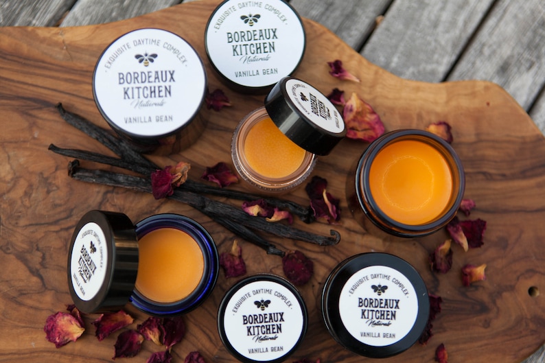 K&ouml;nnte beinhalten: F&uuml;nf kleine Glasgef&auml;&szlig;e mit schwarzen Deckeln, die einen gelben Balsam enthalten. Die Gef&auml;&szlig;e sind auf einer Holzoberfl&auml;che angeordnet, mit Vanilleschoten und Rosenbl&uuml;ten, die um sie herum verstreut sind. Die Etiketten auf den Gef&auml;&szlig;en lauten "EXQUISITE DAYTIME COMPLEX. BORDEAUX KITCHEN Natural VANILLA BEAN."