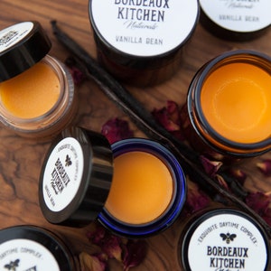 K&ouml;nnte beinhalten: F&uuml;nf kleine Gl&auml;ser Lippenbalsam mit einem blauen, schwarzen und wei&szlig;en Etikett mit der Aufschrift "EXQUISITE DAYTIME COMPLEX. BORDEAUX KITCHEN Naturals VANILLA BEAN". Die Gl&auml;ser stehen auf einer Holzoberfl&auml;che mit getrockneten Rosenbl&uuml;ten und Vanilleschoten.