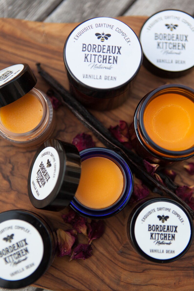 K&ouml;nnte beinhalten: F&uuml;nf kleine Gl&auml;ser Lippenbalsam mit einem blauen, schwarzen und wei&szlig;en Etikett mit der Aufschrift "EXQUISITE DAYTIME COMPLEX. BORDEAUX KITCHEN Naturals VANILLA BEAN". Die Gl&auml;ser stehen auf einer Holzoberfl&auml;che mit getrockneten Rosenbl&uuml;ten und Vanilleschoten.