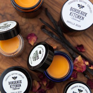 K&ouml;nnte beinhalten: Vier kleine Glasgef&auml;&szlig;e mit Deckeln, die einen gelben Balsam enthalten. Die Gl&auml;ser sind mit "Bordeaux Kitchen Naturals Vanilla Bean" und "Exquisite Daytime Complex" beschriftet. Die Gl&auml;ser befinden sich auf einer Holzoberfl&auml;che mit getrockneten Rosenbl&uuml;ten und Vanilleschoten.