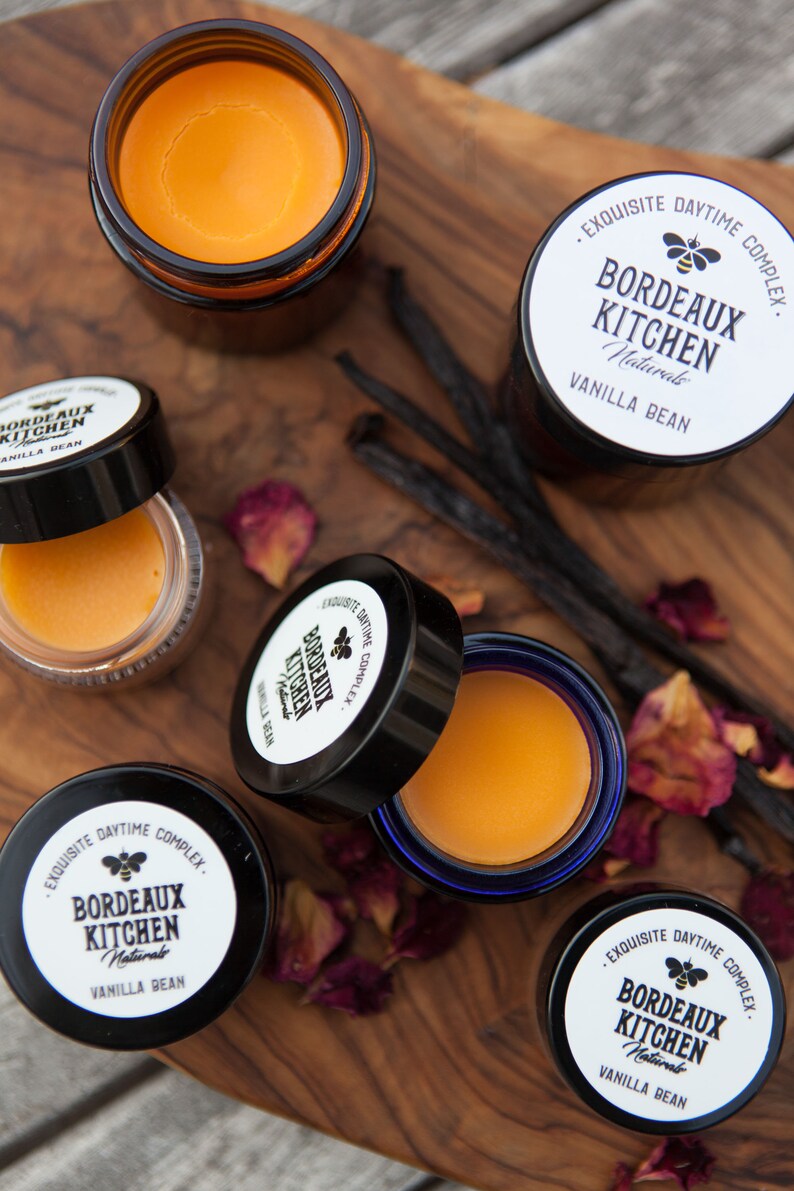 K&ouml;nnte beinhalten: Vier kleine Glasgef&auml;&szlig;e mit Deckeln, die einen gelben Balsam enthalten. Die Gl&auml;ser sind mit "Bordeaux Kitchen Naturals Vanilla Bean" und "Exquisite Daytime Complex" beschriftet. Die Gl&auml;ser befinden sich auf einer Holzoberfl&auml;che mit getrockneten Rosenbl&uuml;ten und Vanilleschoten.