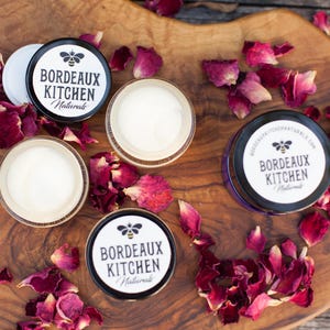 Peut inclure: Trois petits pots en verre contenant de la crème blanche. Chaque pot porte une étiquette noire et blanche sur laquelle est inscrit "Bordeaux Kitchen Naturals". Les pots sont posés sur une surface en bois avec des pétales de rose séchés.