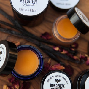 K&ouml;nnte beinhalten: Vier kleine Gl&auml;ser mit Lippenbalsam auf einer Holzoberfl&auml;che mit Vanilleschoten und Rosenbl&uuml;ten. Die Gl&auml;ser sind mit "Bordeaux Kitchen Naturals Vanilla Bean" beschriftet und zeigen ein Bienenlogo.